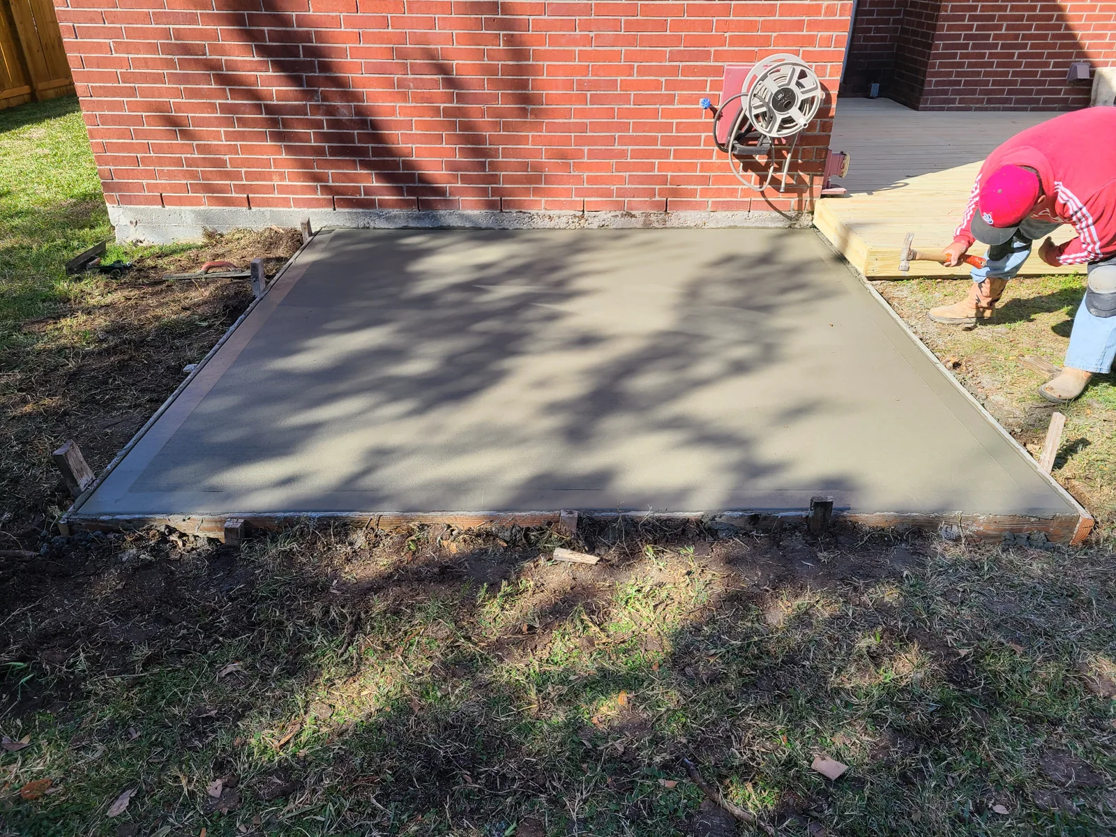 Fresh rectangular concrete patio beside a wooden deck with a crew member finishing the edge, photographed January 2024